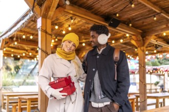 Young diverse couple walking together at a christmas market or winter fair, holding red gift boxes