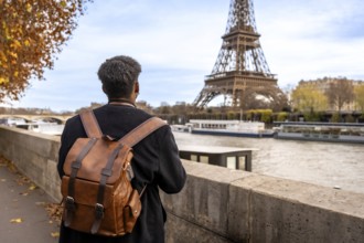 Person with travel backpack standing by the seine, gazing at the eiffel tower amid autumn foliage