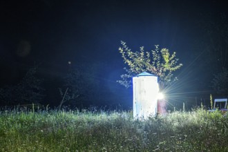 A brightly lit light tent Lichtfang stands in the middle of a traditional orchard next to trees,