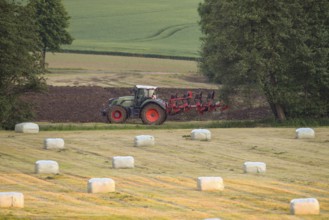 A tractor plows a field in a rural landscape with round bales of hay bales in the foreground,