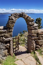 Stone archway with wooden gate in peaceful surroundings, with a view of vast blue lake and sky,