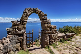 Picturesque stone arch with wooden gate opens to a calm, deep blue lake under clear skies, stone