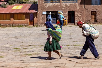Two woman in traditional clothing carry bundles on their backs through a village in Bolivia in