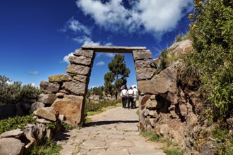 People wearing traditional clothes walk under a rectangular stone arch on a rural path, stone gates