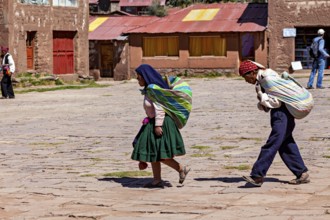 Two woman in colorful clothes carry loads on their backs and walk through a sunny village in