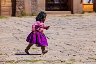 Little girl in purple skirt and pink sweater walks across a paved square, children from Taquile