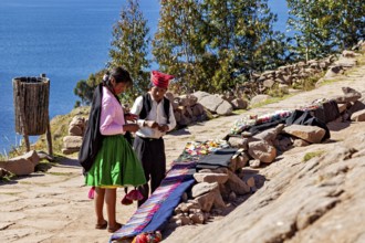 People wearing traditional clothing sell goods on a rock path, overlooking a large lake, the woman