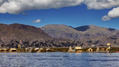 Reed huts on the shores of a tranquil lake in front of a vast mountain landscape under a blue sky,