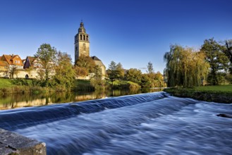 A church on the river with a small waterfall in the foreground and autumn trees in the background,