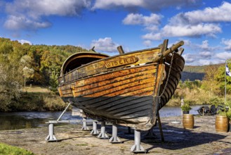 Historic wooden boat on the riverbank on supports, surrounded by autumn trees under a blue sky, the