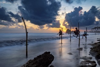 Silhouette of fishermen at sunset over the blue and orange sea, Koggala beach with stilt fishermen