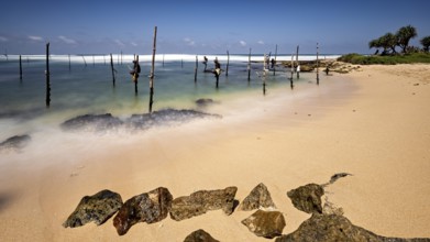 Clear beach with visible rocks and wooden posts in the water, Koggala beach with stilt fishermen in