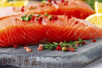 Two pieces of salmon fillet sit on a wooden cutting board. They are topped with herbs and pink