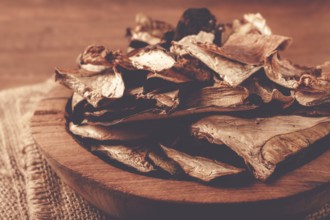 A collection of dried mushrooms fills a wooden bowl placed on a cloth surface. The mushrooms are
