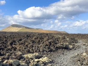 Hiking trail through lava rock to Caldera Blanca volcano against blue sky with white clouds, Mancha