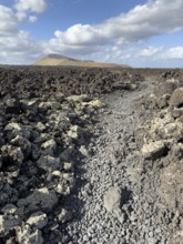 Hiking trail through lava rock to Caldera Blanca volcano against blue sky with white clouds, Mancha