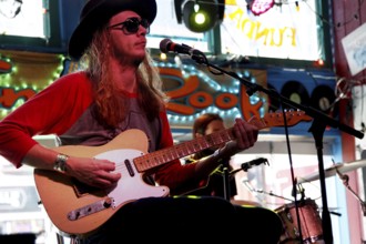 Musician wearing hat and sunglasses, playing guitar on stage in front of colorful window