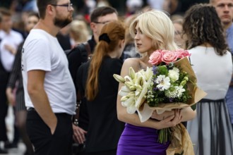 Blonde woman holding a bouquet of flowers during a high school graduation ceremony in a busy