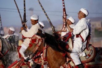 Group of riders wearing traditional clothing hold up rifles at a historic Fantasia event, El Kelâa