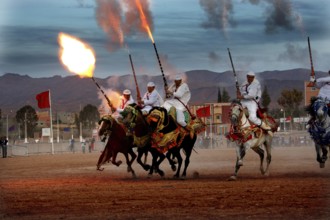 Riders on horses shoot volleys synchronously into the evening sky at Fantasia, El Kelâa m'Gouna,