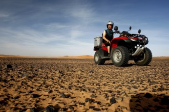 A red quad bike rides across a rocky desert landscape under a blue sky, Erg Chebbi, Hammada,