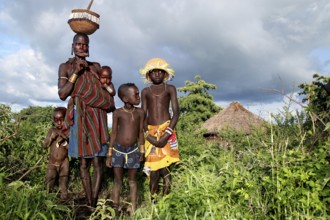 Mursi woman with children in front of a grass hut and cloudy sky, Mago National Park, Ethiopia