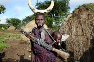 Man with gun and child on his back, straw huts in the background in Mago National Park, Mago