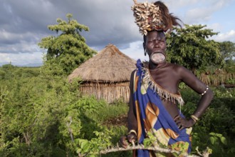 Mursi woman with lip plate in front of a hut in Mago National Park. The landscape is green and the