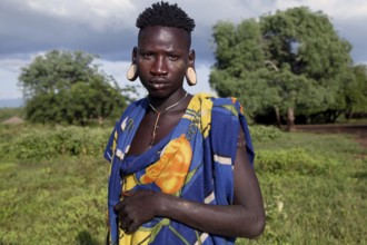 Mursi man wearing traditional clothing and earrings in front of lush landscape in Mago National