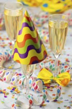 Three champagne glasses, one decorated with a colorful party hat, surrounded by cheerful confetti