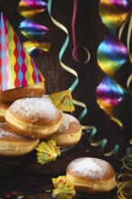 A pile of donuts with powdered sugar, colorful garlands and party hat against a dark wooden