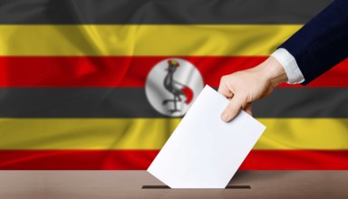 Uganda presidential vote 2026. Hand placing a ballot in ballot box with the Ugandan flag in the