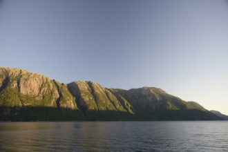 Rocky mountain range along a body of water under clear sky at Lysefjord at sunrise, Lysefjord,