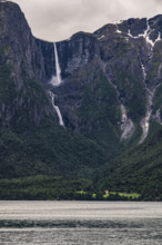 A narrow waterfall plunges from dramatic mountains into a dark fjord