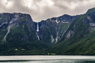 A waterfall flows between steep mountains into a quiet fjord under a cloudy sky