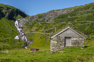 A stone hut stands in front of a rippling waterfall in the middle of green meadows