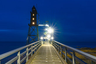 An illuminated lighthouse at night that can be reached via a bridge with bright lights
