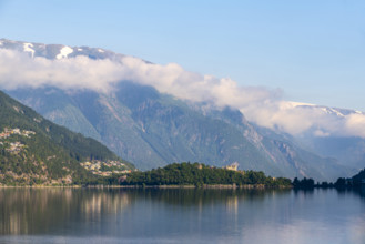 A calm lake reflects the mountains covered by clouds and the morning light