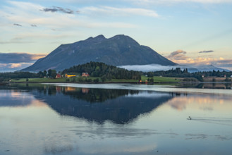 A mountain is reflected in a peaceful lake, surrounded by meadows and fields under an evening sky