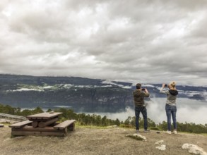 Utvik, Vestland, Norway, Two people taking pictures of a misty mountain backdrop with a lake in the