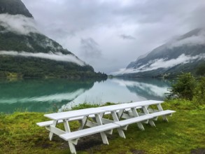 Olden, Vestland, Norway, white picnic table at a lake with mountains and low-hanging clouds in the