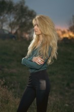 A woman stands with her arms crossed on a grassy hill. She faces the horizon where the sun is