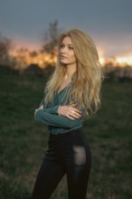 A woman poses with crossed arms while standing on grass. She has long hair and a casual outfit. The