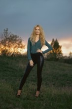 A woman poses on a grassy hill at sunset. The sky shows shades of orange and purple. Trees are in