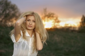A woman stands in a field with blonde hair flowing as she poses during sunset. The sun creates a