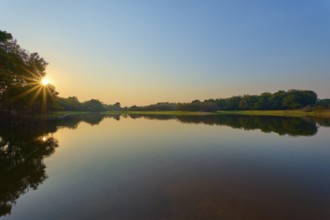 The first rays of sunshine fall on the calm lake, surrounded by green trees and clear skies,