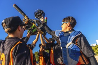 Young baseball players huddling together, lifting catcher's gear and a bat into the bright blue
