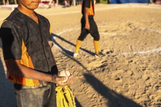 Young baseball boy in orange and black uniform holds glove and ball on sunlit dirt field during
