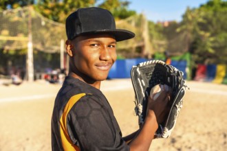 Young male baseball player stands on a sunny field, smiling confidently while holding a baseball in