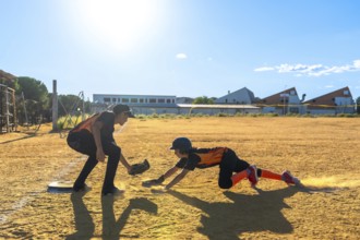 Kids playing baseball on a dusty field, one player sliding into home plate kicking up dust while